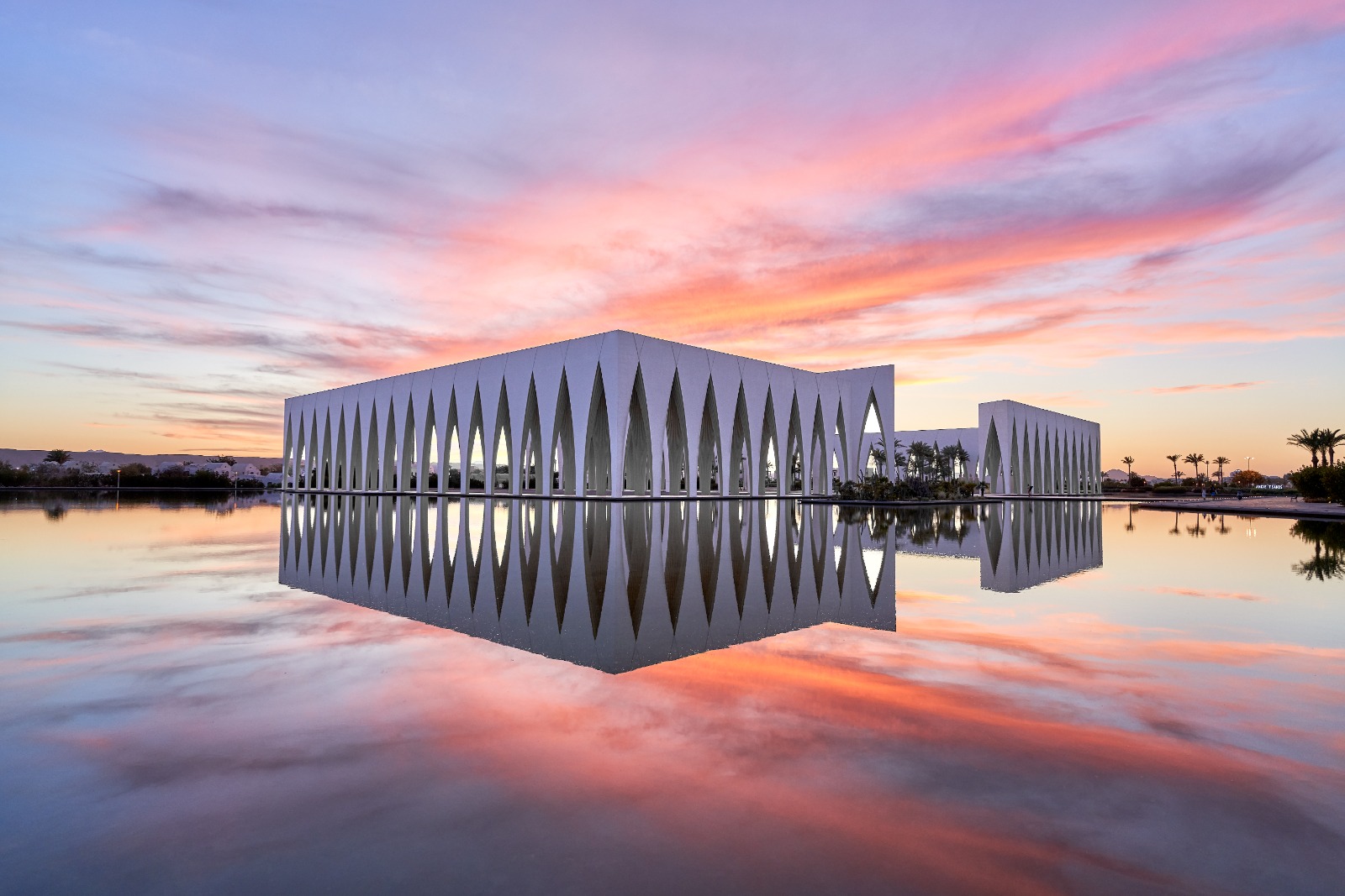 The Gouna Conference and Culture Center at sunset, its white modern architecture perfectly reflected in the calm water of a lagoon under a pink and orange sky.